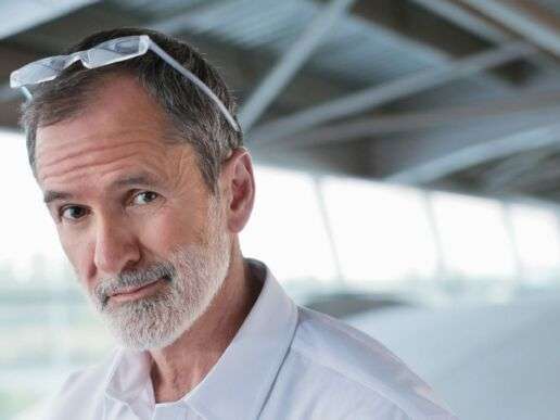Älterer Herr mit stylischer Brille guckt verschmitzt in die Kamera, im Hintergrund eine Bedachung mit Ausblick