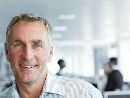 Mitarbeiter innerhalb eines Büros schaut freundlich in die Kamera, während hinter ihm eine Besprechung abgehalten wird