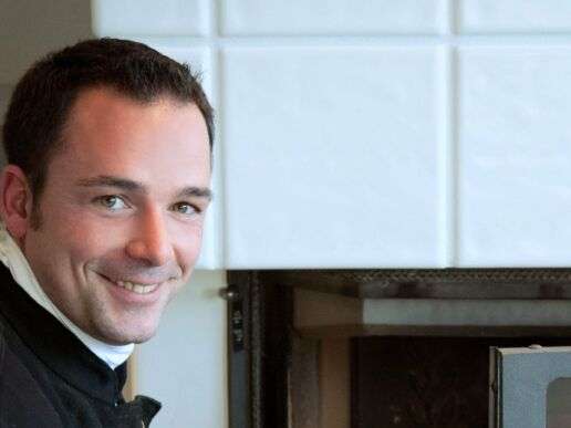 Schornsteinfeger vor einem brennendem Kamin lächelt freundlich in die Kamera